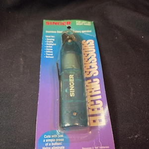 May include: A Singer brand battery-operated electric scissor tool in a teal plastic casing, packaged on a blue and purple cardboard backing. The packaging features the text "ELECTRIC SCISSORS," "SINGER," and "Requires 2 AA batteries (not included)."