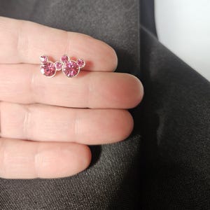 Op de afbeelding: Een paar zilverkleurige oorstekers met een Mickey Mouse-ontwerp. Elke oorbel heeft drie roze, sprankelende, rond geslepen edelstenen. De oorbellen worden gepresenteerd tegen een donkergrijze achtergrond.