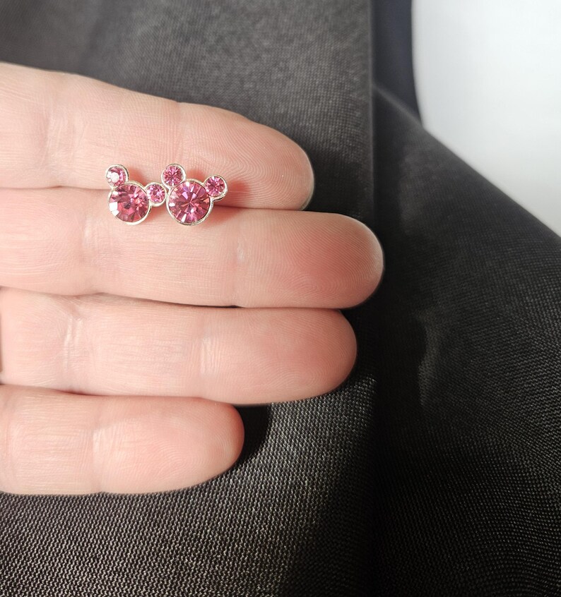 Op de afbeelding: Een paar zilverkleurige oorstekers met een Mickey Mouse-ontwerp. Elke oorbel heeft drie roze, sprankelende, rond geslepen edelstenen. De oorbellen worden gepresenteerd tegen een donkergrijze achtergrond.