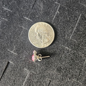 Op de afbeelding: Een zilveren oorbel met een roze edelsteen, naast een Amerikaanse kwart dollar munt. De oorbel heeft een pin en vlindersluiting. De edelsteen heeft een vierkante vorm met een puntige top.