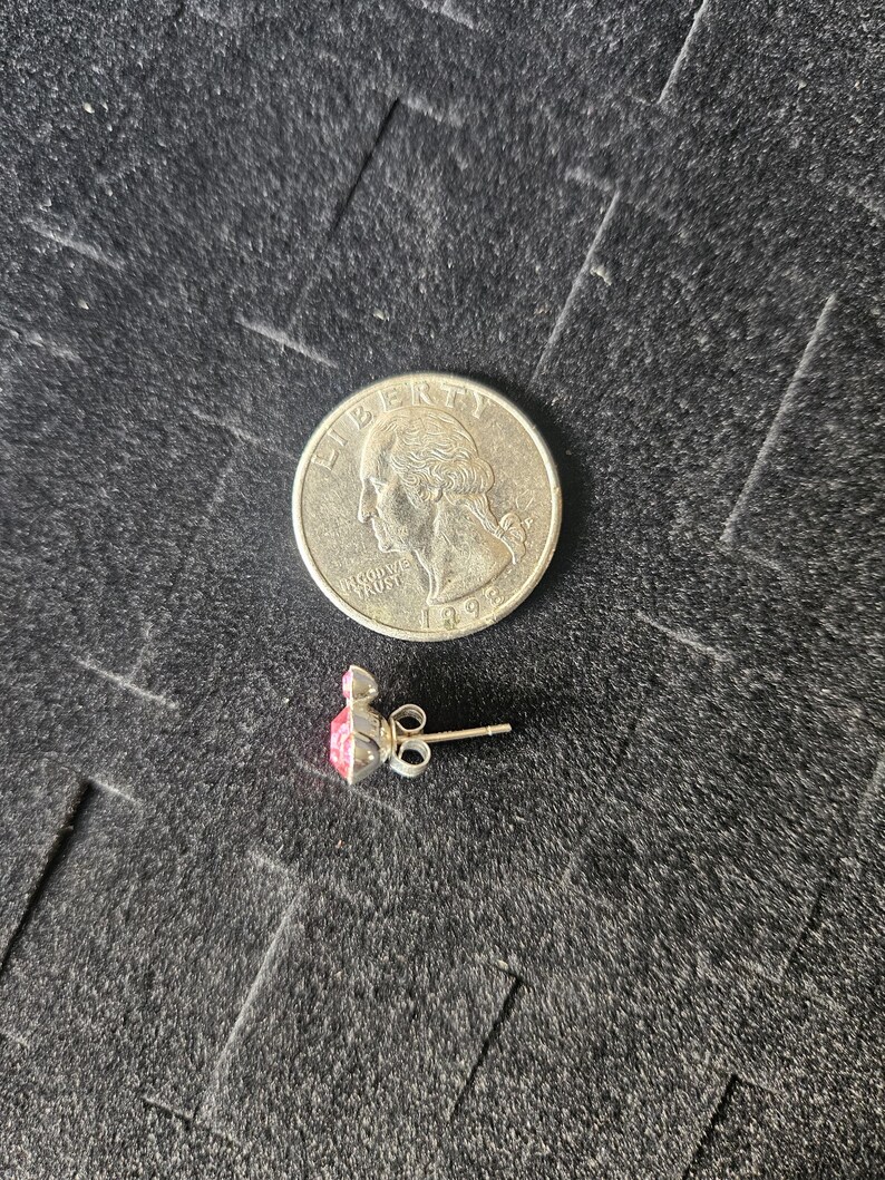 Op de afbeelding: Een zilveren oorbel met een roze edelsteen, naast een Amerikaanse kwart dollar munt. De oorbel heeft een pin en vlindersluiting. De edelsteen heeft een vierkante vorm met een puntige top.