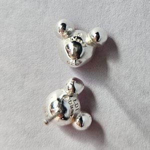 Op de afbeelding: Twee zilverkleurige oorstekers in de vorm van Mickey Mouses hoofd. Elke oorbel heeft drie afgeronde vormen, waarvan de middelste iets groter is. De oorbellen liggen op een lichtgrijze ondergrond.