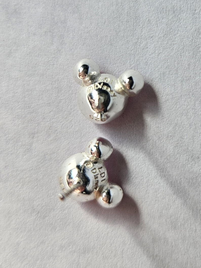 Op de afbeelding: Twee zilverkleurige oorstekers in de vorm van Mickey Mouses hoofd. Elke oorbel heeft drie afgeronde vormen, waarvan de middelste iets groter is. De oorbellen liggen op een lichtgrijze ondergrond.