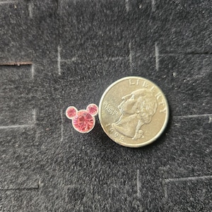 Op de afbeelding: Een kleine, roze, met strass-steentjes bezette bedel in de vorm van Mickey Mouses hoofd, naast een Amerikaanse kwart dollar. De bedel heeft drie cirkelvormige elementen, twee voor de oren en &eacute;&eacute;n voor het hoofd, allemaal gevuld met roze strass-steentjes. De munt is zilverkleurig.