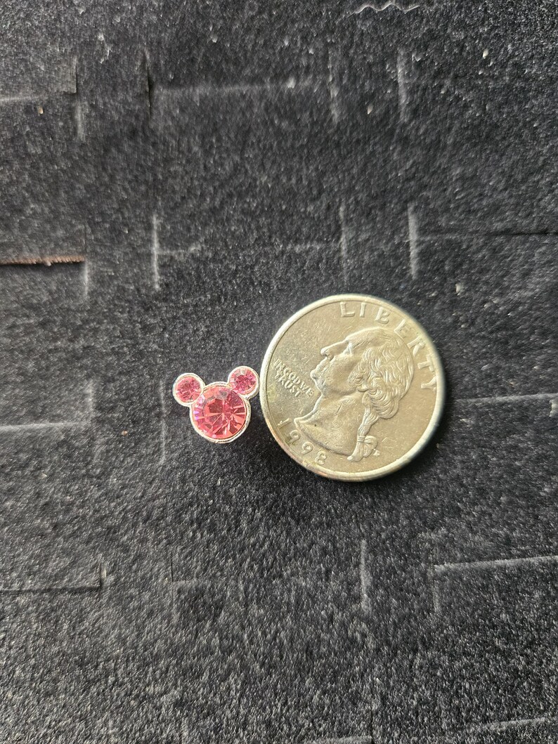 Op de afbeelding: Een kleine, roze, met strass-steentjes bezette bedel in de vorm van Mickey Mouses hoofd, naast een Amerikaanse kwart dollar. De bedel heeft drie cirkelvormige elementen, twee voor de oren en &eacute;&eacute;n voor het hoofd, allemaal gevuld met roze strass-steentjes. De munt is zilverkleurig.