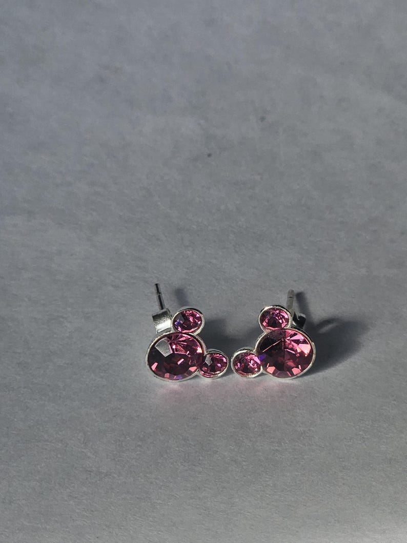 Op de afbeelding: Een paar oorstekers met een Mickey Mouse-ontwerp. Elke oorbel heeft een zilverkleurige metalen achterkant en is versierd met drie roze, gefacetteerde edelstenen. De oorbellen zijn geplaatst tegen een lichtgrijze achtergrond.