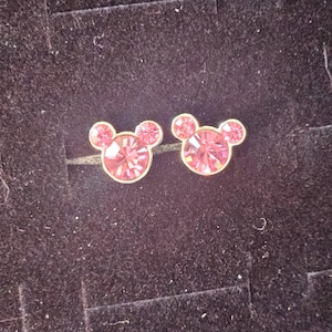 Op de afbeelding: Een paar roze kristallen oorstekers in de vorm van Mickey Mouse. De oorbellen hebben een goudkleurige zetting en worden gepresenteerd op een donkere, getextureerde ondergrond.