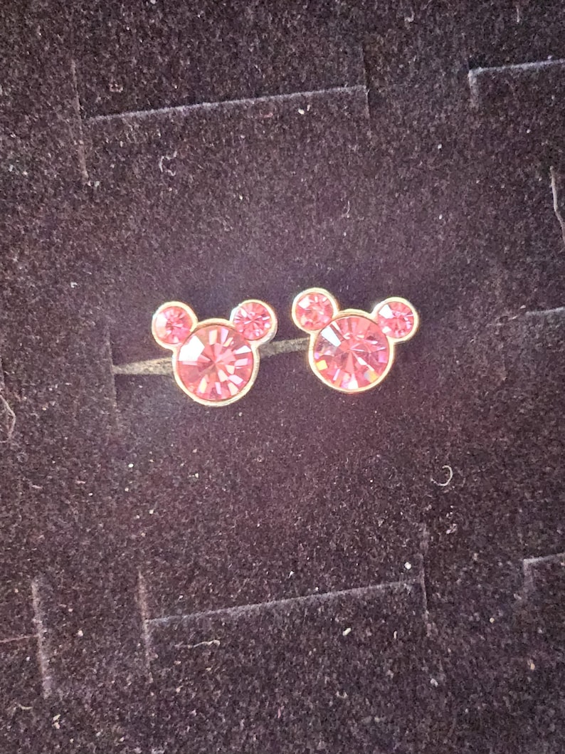 Op de afbeelding: Een paar roze kristallen oorstekers in de vorm van Mickey Mouse. De oorbellen hebben een goudkleurige zetting en worden gepresenteerd op een donkere, getextureerde ondergrond.