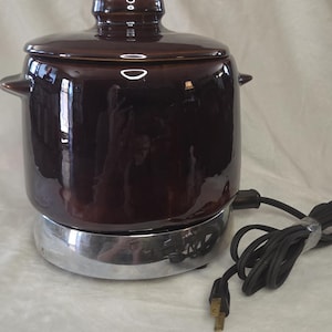 May include: A dark brown ceramic slow cooker with a matching lid and small side handles. The pot sits on a circular silver-colored metal base with a black electrical cord and a two-prong plug extending from the side of the unit.