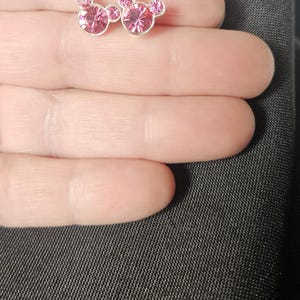 Op de afbeelding: Een paar oorstekers met een Mickey Mouse-ontwerp. Elke oorbel heeft een zilverkleurige zetting met een roze, edelsteenachtig middenstuk en kleinere roze edelstenen voor de oren. De oorbellen worden gepresenteerd op een lichte huidskleur achtergrond.