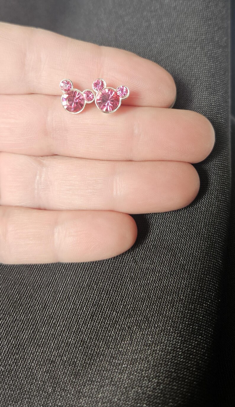 Op de afbeelding: Een paar oorstekers met een Mickey Mouse-ontwerp. Elke oorbel heeft een zilverkleurige zetting met een roze, edelsteenachtig middenstuk en kleinere roze edelstenen voor de oren. De oorbellen worden gepresenteerd op een lichte huidskleur achtergrond.