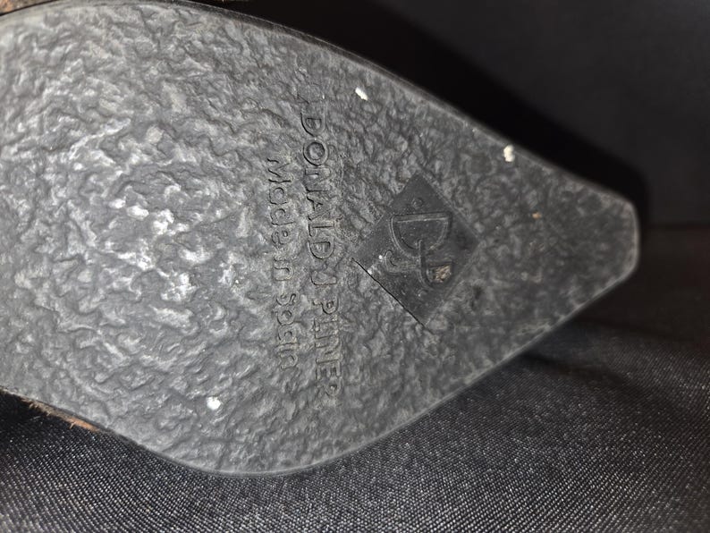 May include: Close-up of a black shoe sole with a textured surface. The sole has the brand name "DONALD J. PLINER" and "made in Spain" printed on it, along with a logo.