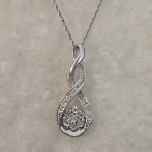 May include: A silver pendant necklace featuring an infinity symbol design. The pendant is adorned with small, sparkling gemstones and a cluster of larger stones at the center. The necklace hangs on a delicate chain.