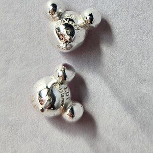 Op de afbeelding: Twee zilverkleurige Mickey Mouse bedel kralen. Elke kraal heeft het iconische ontwerp met drie cirkels, waarbij de centrale cirkel iets groter is. De kralen hebben een glanzend, reflecterend oppervlak.