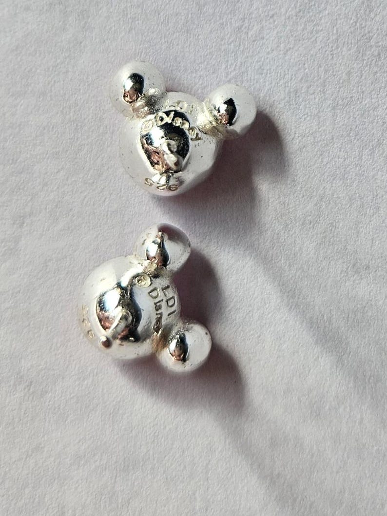 Op de afbeelding: Twee zilverkleurige Mickey Mouse bedel kralen. Elke kraal heeft het iconische ontwerp met drie cirkels, waarbij de centrale cirkel iets groter is. De kralen hebben een glanzend, reflecterend oppervlak.