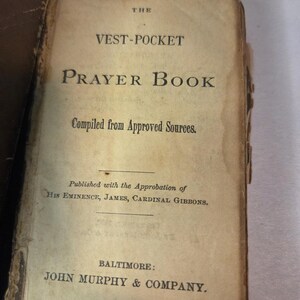 May include: An antique, small-format book titled The Vest-Pocket Prayer Book, Compiled from Approved Sources. The title page features black serif text on aged, off-white paper, noting publication by John Murphy & Company in Baltimore.