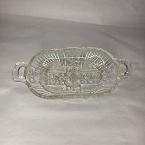 May include: A clear, oval-shaped glass dish featuring two integrated handles on the sides. The surface is decorated with an embossed floral pattern in the center and radiating fan-like lines along the edges, creating a textured appearance.