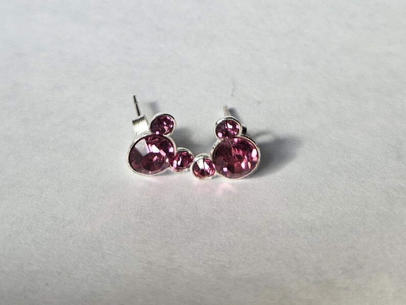 Op de afbeelding: Een paar oorstekers met een ontwerp van drie cirkels met roze edelstenen. De oorbellen hebben een zilverkleurige metalen zetting en een steker sluiting. De edelstenen hebben een levendige roze kleur.