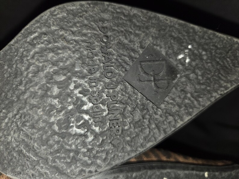 May include: Close-up of a shoe sole with a textured, dark gray surface. The words "DONALD J PLINER made in Spain" are embossed on the sole. A black square with the letters "DJ" is also visible.