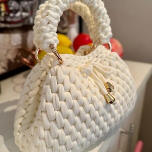 May include: Cream-colored, hand-knitted handbag with a woven texture. The bag features a rounded shape, a thick, braided handle, and gold-colored metal hardware. A drawstring closure with gold accents secures the top.