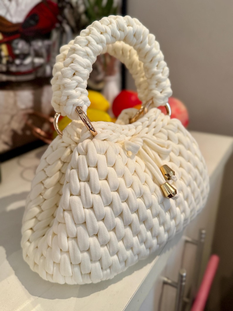 May include: Cream-colored, hand-knitted handbag with a woven texture. The bag features a rounded shape, a thick, braided handle, and gold-colored metal hardware. A drawstring closure with gold accents secures the top.