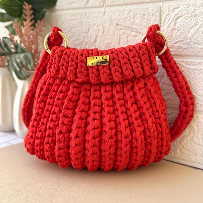 Pode incluir: Bolsa de croch&ecirc; vermelha vibrante com al&ccedil;as de anel de metal dourado. A bolsa apresenta uma etiqueta retangular dourada com o texto "TIFE & CRO". O corpo da bolsa tem uma textura canelada e as al&ccedil;as s&atilde;o tran&ccedil;adas.
