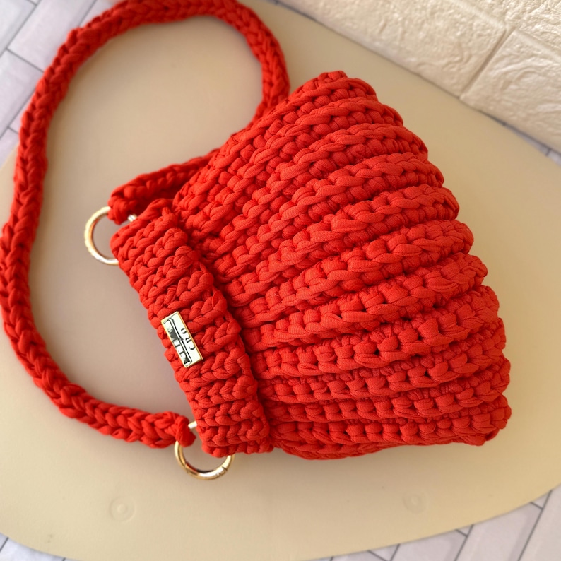 Pode incluir: Bolsa de croch&ecirc; vermelha brilhante, feita &agrave; m&atilde;o, com al&ccedil;a tran&ccedil;ada e argolas de metal dourado. A bolsa apresenta uma etiqueta retangular com o texto "CRO". A bolsa &eacute; feita de fio grosso e texturizado.