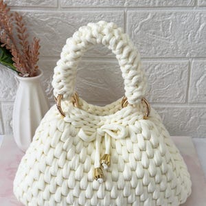 Può includere: Borsa a mano color crema, lavorata all'uncinetto a mano, di forma arrotondata e con un manico spesso ad anello. La borsa presenta anelli metallici color oro e chiusura con coulisse con dettagli dorati. La borsa è realizzata in filato morbido e strutturato.