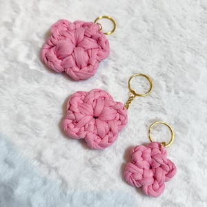 Könnte beinhalten: Drei rosa, blütenförmige Schlüsselanhänger mit goldfarbenen Schlüsselringen. Die Schlüsselanhänger sind aus gewebtem Stoff gefertigt und die Blüten haben ein strukturiertes Aussehen. Die Schlüsselanhänger werden auf einer weißen, flauschigen Oberfläche präsentiert.