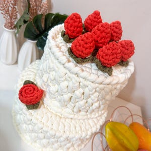 May include: A white crocheted hat with a brim, adorned with red crocheted strawberries and a red rose. The strawberries have green leaves. The hat is handmade and has a textured appearance.