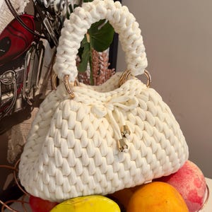 May include: Cream-colored, hand-knitted handbag with a rounded shape and a thick, braided handle. The bag features gold-colored metal rings and a drawstring closure. The bag is sitting on top of a wire basket filled with fruit.