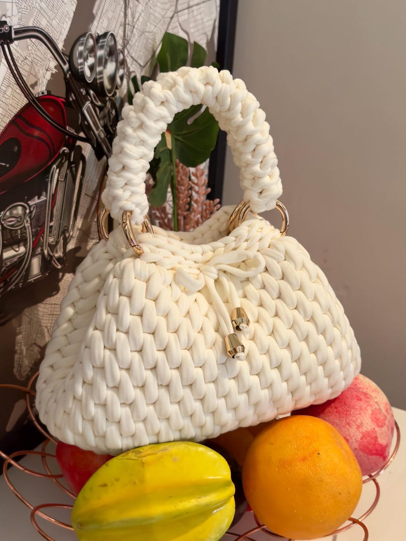 May include: Cream-colored, hand-knitted handbag with a rounded shape and a thick, braided handle. The bag features gold-colored metal rings and a drawstring closure. The bag is sitting on top of a wire basket filled with fruit.