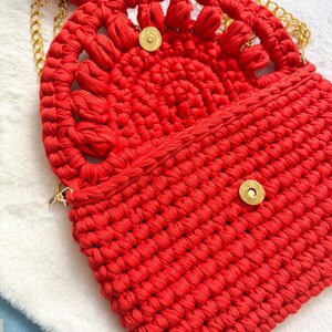 May include: A bright red, handmade crochet bag with a gold chain strap. The bag features a decorative, openwork design on the flap and a tassel. The bag is secured with a gold magnetic snap closure.