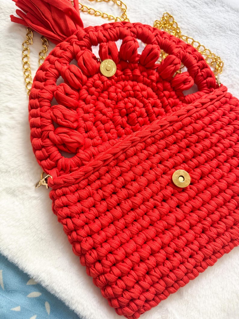 May include: A bright red, handmade crochet bag with a gold chain strap. The bag features a decorative, openwork design on the flap and a tassel. The bag is secured with a gold magnetic snap closure.