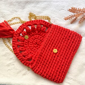May include: A red crochet handbag with a gold chain strap and tassel. The bag features a circular design with a floral pattern on the front flap and a rectangular body. The bag is secured with a gold magnetic snap.
