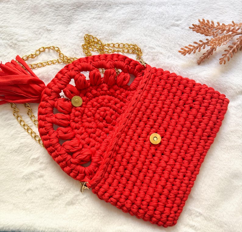 May include: A red crochet handbag with a gold chain strap and tassel. The bag features a circular design with a floral pattern on the front flap and a rectangular body. The bag is secured with a gold magnetic snap.