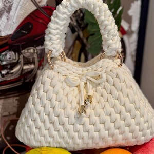 May include: Cream-colored, hand-knitted handbag with a rounded shape and a braided handle. The bag features gold-colored metal rings and decorative tassels. The bag's texture is prominent, showcasing the knitted pattern.