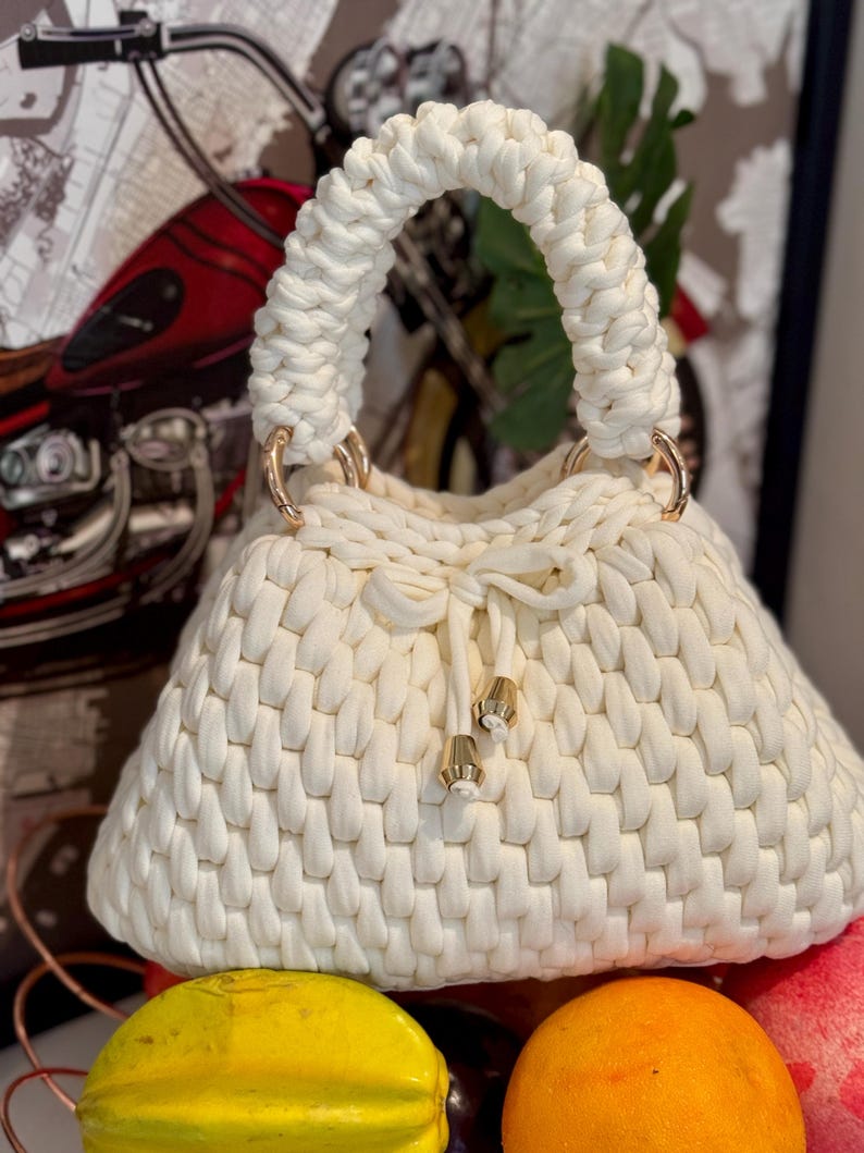 May include: Cream-colored, hand-knitted handbag with a rounded shape and a braided handle. The bag features gold-colored metal rings and decorative tassels. The bag's texture is prominent, showcasing the knitted pattern.