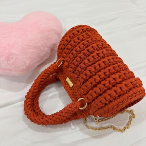 May include: A handmade, burnt orange crochet handbag with a cylindrical shape. The bag features two handles, gold-colored metal rings, and a gold chain strap. A pink heart-shaped pillow is in the background.