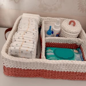 Pode incluir: Um cesto de fraldas de crochê branco e coral com vários compartimentos. O cesto contém fraldas, lenços umedecidos para bebês, loção para bebês e um pequeno recipiente com tampa coral. O cesto foi projetado para guardar itens essenciais para bebês.