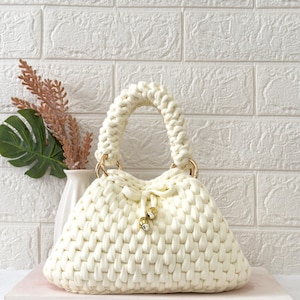 May include: Cream-colored, hand-crocheted handbag with a rounded shape and a braided handle. The bag features gold-colored metal rings and a drawstring closure with decorative gold-colored beads. The bag is made of a thick, textured yarn.