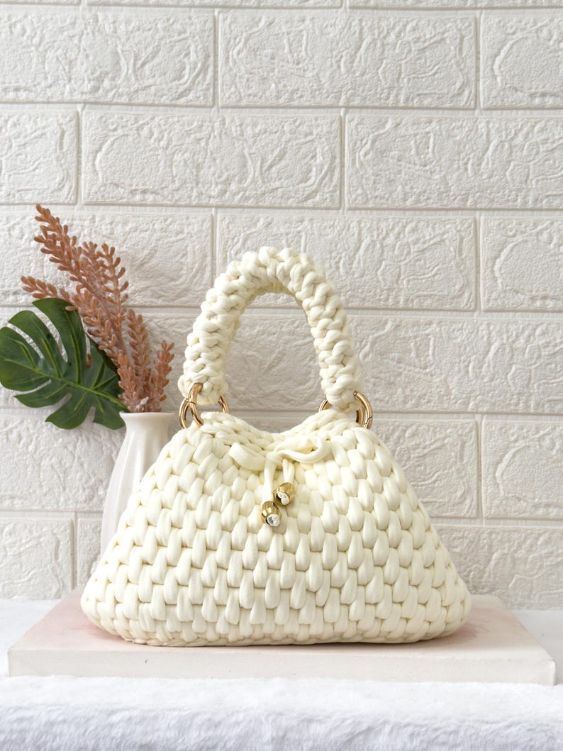 May include: Cream-colored, hand-crocheted handbag with a rounded shape and a braided handle. The bag features gold-colored metal rings and a drawstring closure with decorative gold-colored beads. The bag is made of a thick, textured yarn.