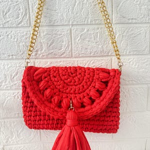 May include: A bright red crochet crossbody bag with a gold chain strap. The bag features a decorative flap with a floral design and a matching tassel. The bag is made of a thick, textured yarn.