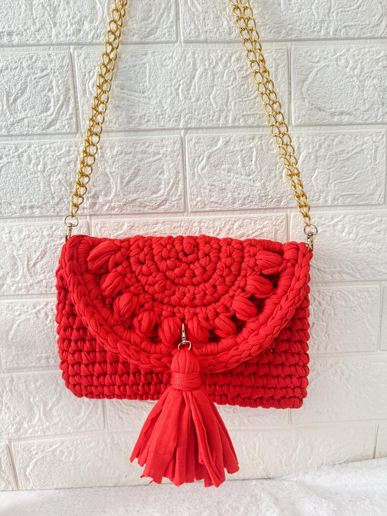 May include: A bright red crochet crossbody bag with a gold chain strap. The bag features a decorative flap with a floral design and a matching tassel. The bag is made of a thick, textured yarn.