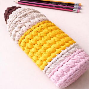 May include: A handmade pencil-shaped crochet case in brown, beige, yellow, and pink. The case is next to three pencils with orange, pink, and purple accents. The case is designed to hold pencils or other writing utensils.