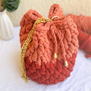 May include: A handmade, crochet bucket bag in shades of orange and red. The bag features a gold chain strap and drawstring closure with gold-colored accents. The bag is made of a thick, textured yarn.