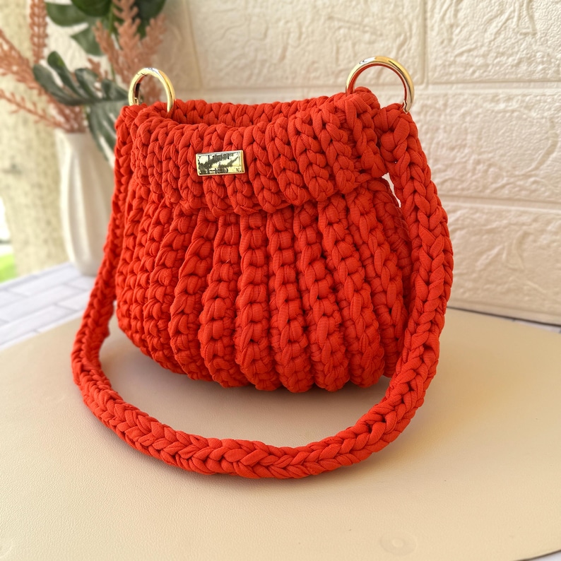 Pode incluir: Uma vibrante bolsa de m&atilde;o vermelha de croch&ecirc; com al&ccedil;as de anel de metal dourado e um pequeno fecho retangular dourado. A bolsa apresenta uma al&ccedil;a tran&ccedil;ada e um design texturizado e canelado. Um acess&oacute;rio elegante.