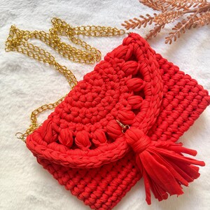 May include: A handmade red crochet clutch with a gold chain strap and a matching tassel. The bag features a textured design and a flap closure. The clutch is a stylish accessory for any occasion.