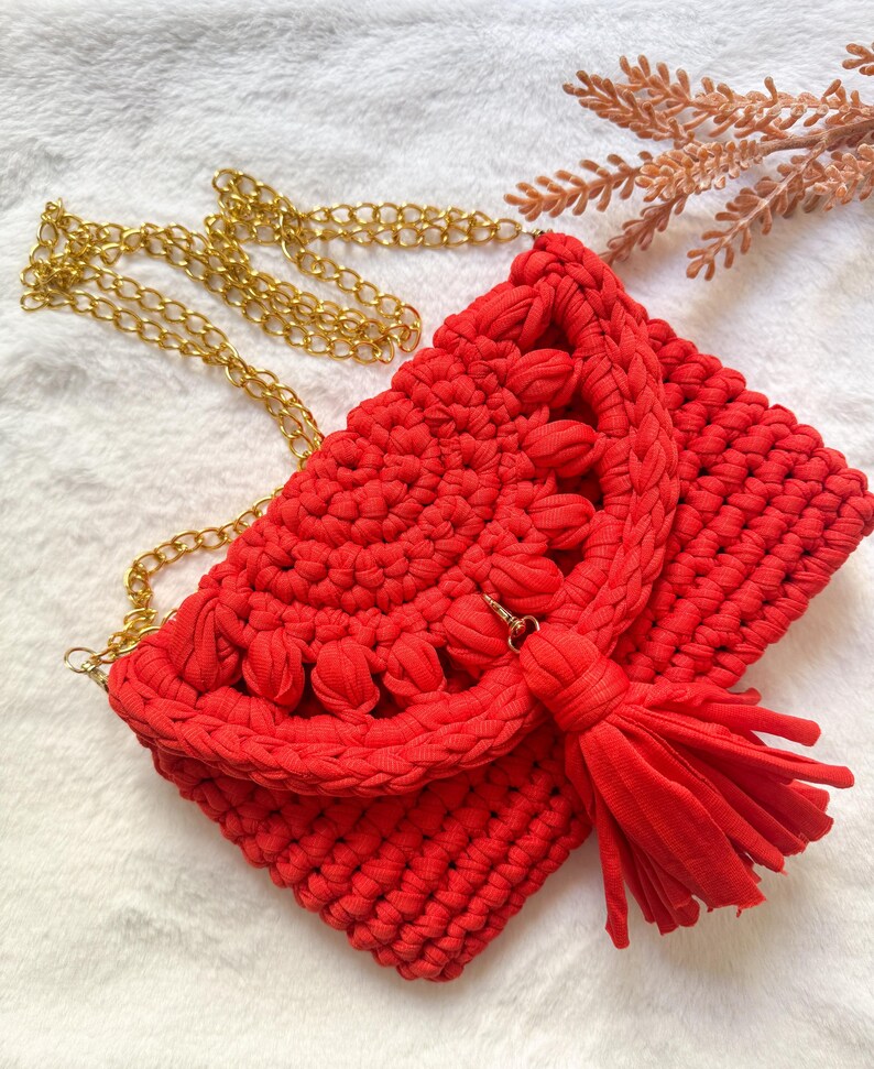 May include: A handmade red crochet clutch with a gold chain strap and a matching tassel. The bag features a textured design and a flap closure. The clutch is a stylish accessory for any occasion.