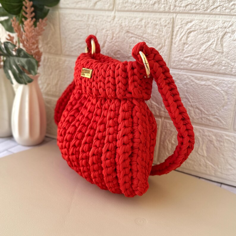 Pode incluir: Uma vibrante bolsa vermelha, feita &agrave; m&atilde;o em croch&ecirc;, com ferragens met&aacute;licas douradas. A bolsa apresenta um formato arredondado, uma al&ccedil;a tran&ccedil;ada e uma pequena etiqueta retangular dourada. A bolsa &eacute; exibida contra uma parede de tijolos brancos.